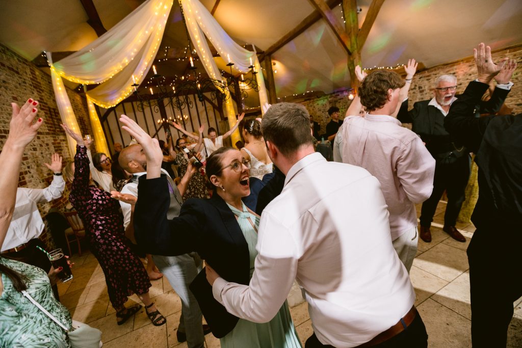 People dancing with raised arms at an indoor event with fairy lights and draped fabric on the ceiling; a festive atmosphere with brick walls and guests in formal attire, perfect for capturing memorable Upwaltham Barns Wedding Photography moments.