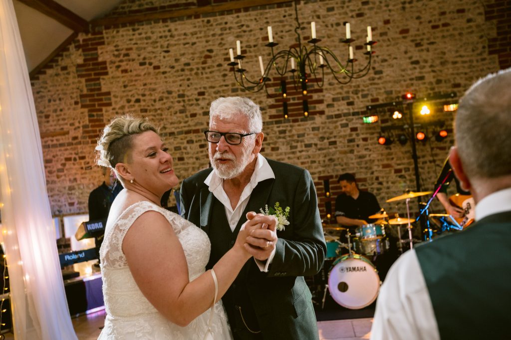 A bride in a white dress dances with an older man in a suit at a wedding reception, with a band and musical instruments visible in the background, beautifully captured by Upwaltham Barns Wedding Photography.