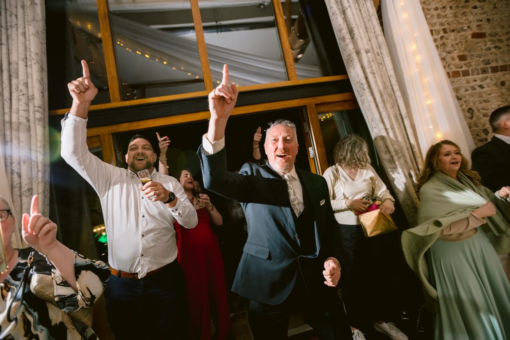 A group of people dressed in formal attire stand indoors at an Upwaltham Barns wedding, smiling and raising one finger in the air as if dancing or celebrating at the event.