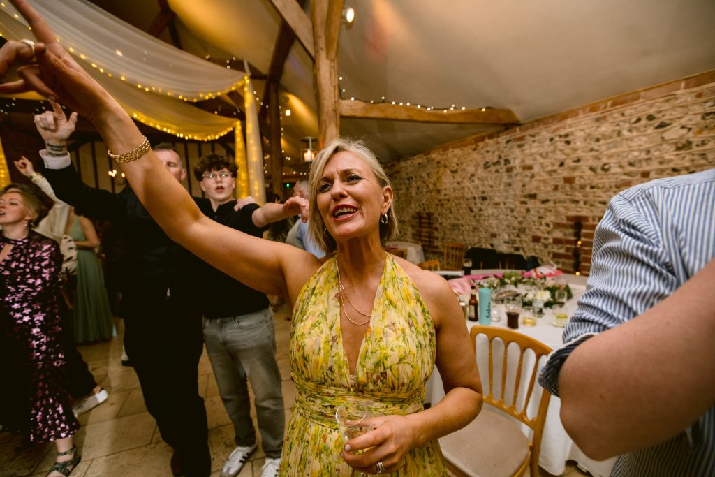 A woman in a yellow dress smiles and dances with her hand raised at an indoor event at Upwaltham Barns, surrounded by guests, fairy lights, and decorated tables—a perfect moment for Upwaltham Barns wedding photography.
