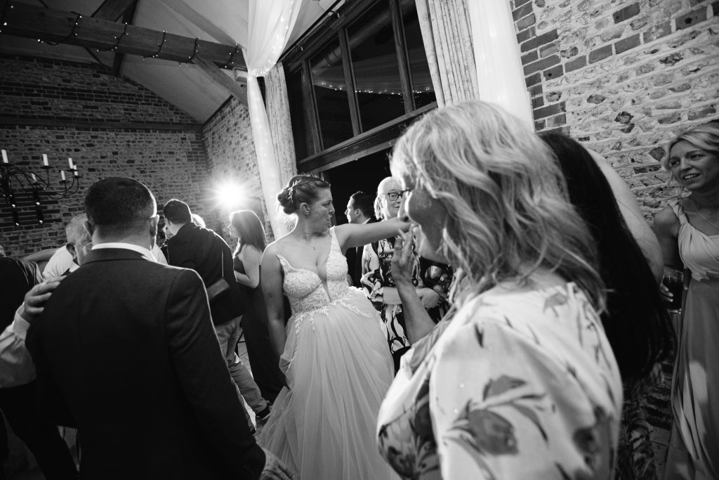 A bride in a wedding dress dances among guests in a decorated indoor venue with brick walls and large windows, capturing the lively and celebratory spirit of Upwaltham Barns wedding photography.
