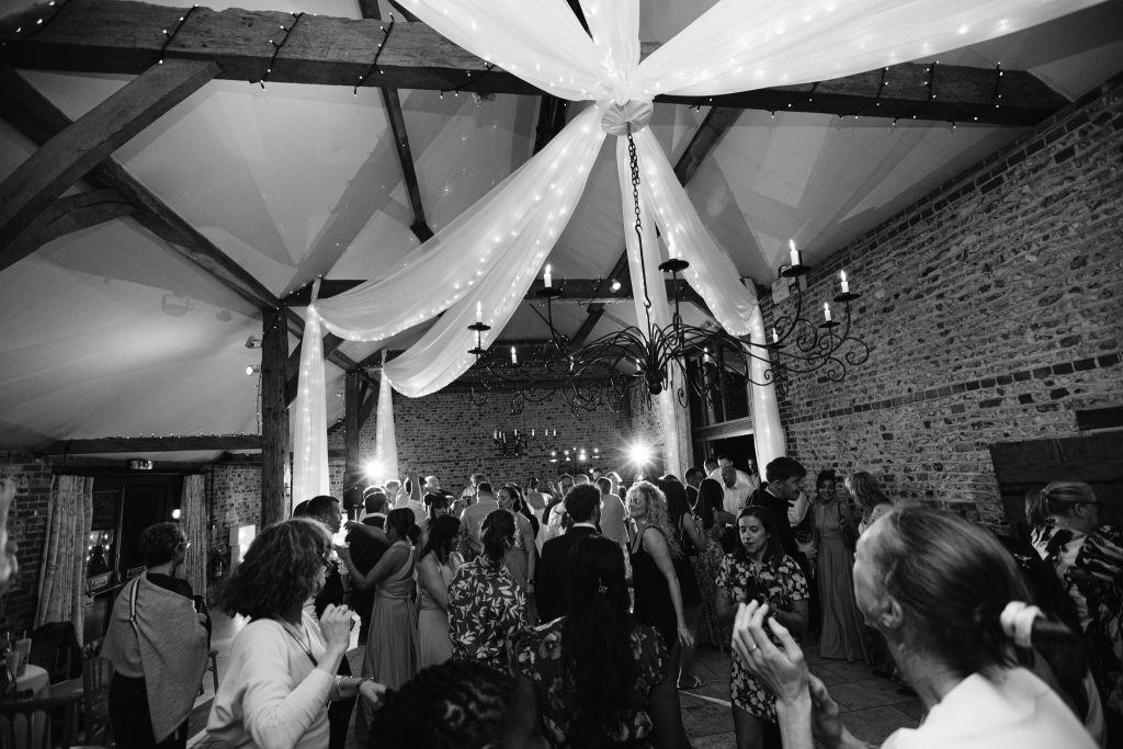 A group of people are dancing and socialising in a decorated indoor venue with draped fabric and exposed brick walls, capturing the lively atmosphere typical of Upwaltham Barns Wedding Photography.