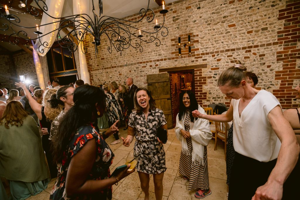 A group of people in semi-formal attire are dancing and laughing together in a rustic indoor venue with exposed brick walls and warm lighting, perfectly capturing the joy of Upwaltham Barns Wedding Photography.