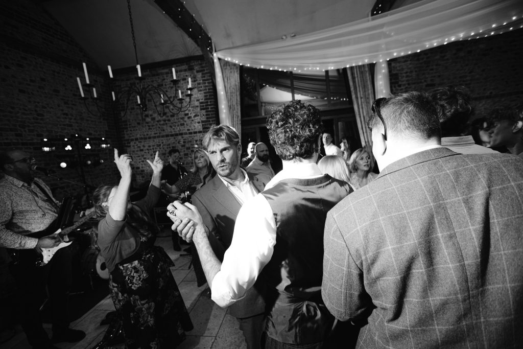 A group of people, some dancing and some talking, gather in a brick-walled venue with fairy lights and a band performing—capturing the lively atmosphere perfect for Upwaltham Barns wedding photography.