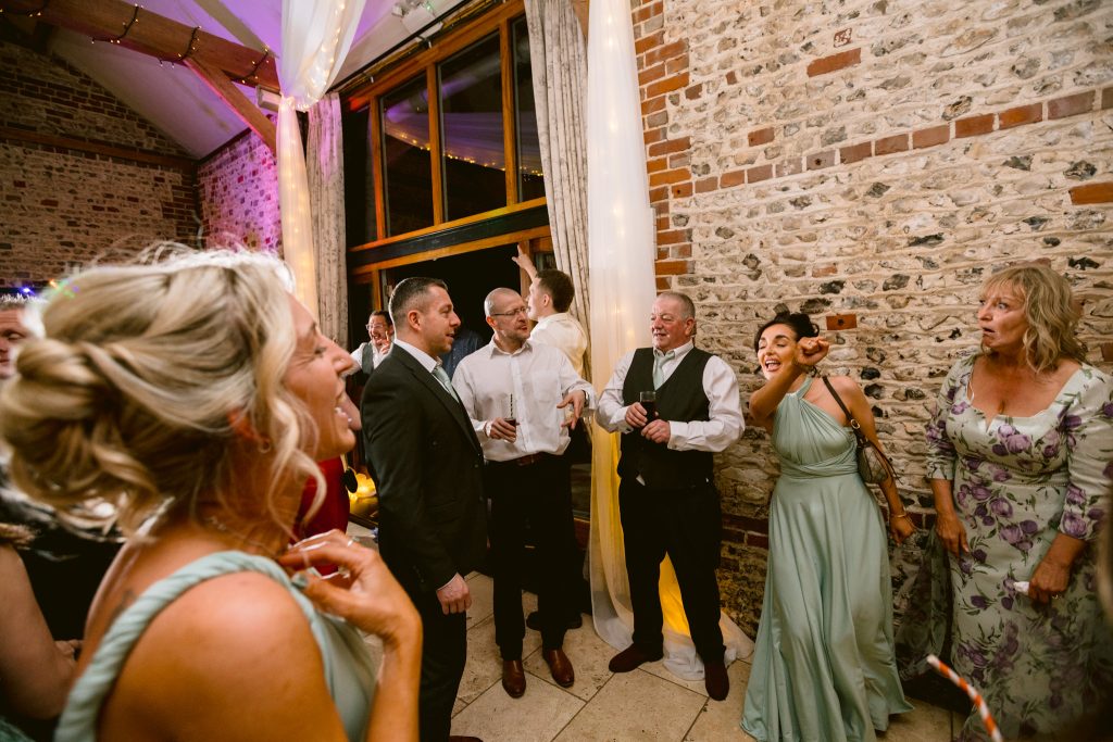 A group of people in formal attire are talking and laughing at an indoor event with brick walls and large windows; one person, captured in this Upwaltham Barns Wedding Photography moment, is adjusting curtains in the background.