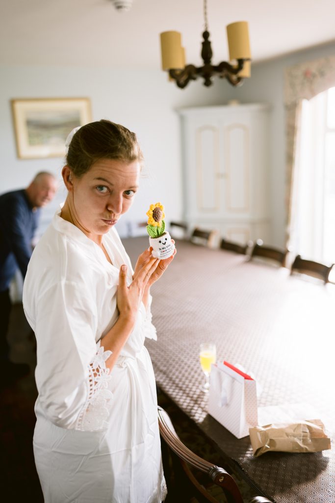 A woman in a white dressing gown stands by a dining table holding a small potted sunflower, while a man lingers in the background—an intimate moment captured in classic Upwaltham Barns Wedding Photography style.