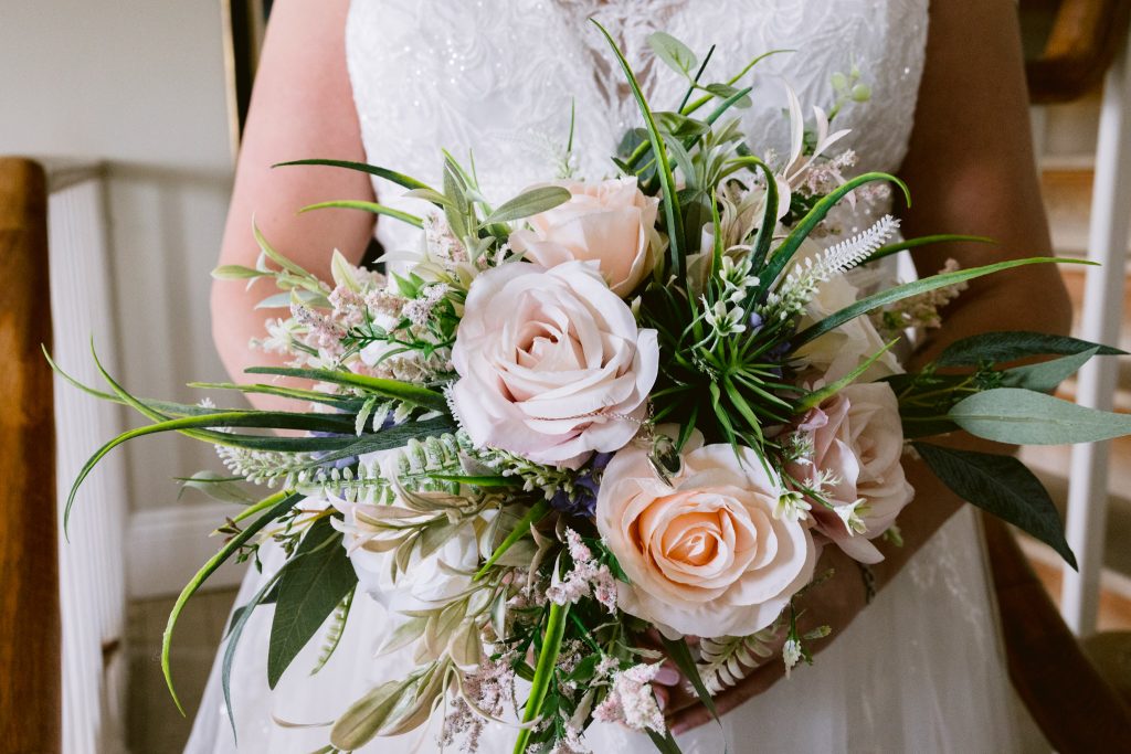 A person in a white lace dress holds a bouquet of pale pink roses and assorted greenery, beautifully captured in this Upwaltham Barns Wedding Photography moment.
