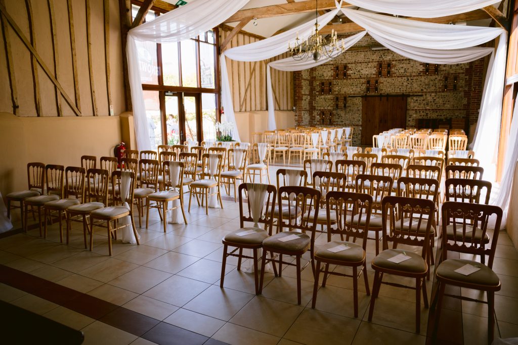 Rows of empty wooden chairs are arranged in a decorated hall at Upwaltham Barns, with white drapes and natural light streaming through large windows, ready for a beautiful wedding ceremony and stunning wedding photography.