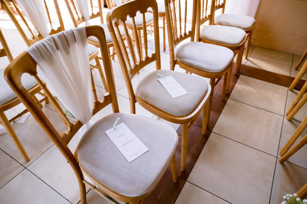 Rows of wooden chairs with white seat cushions and reserved cards are arranged indoors for an event, evoking the timeless charm often captured in Upwaltham Barns Wedding Photography.