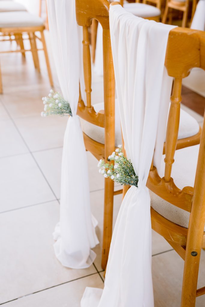 Two wooden chairs with white fabric draped over the backs, tied with small bouquets of white flowers, arranged in a bright indoor setting—an elegant detail perfect for Upwaltham Barns Wedding Photography.
