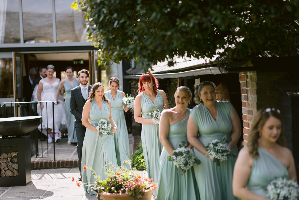 A group of bridesmaids in light blue dresses walk ahead of a bride and groom exiting a building during an outdoor Upwaltham Barns Wedding Photography procession.