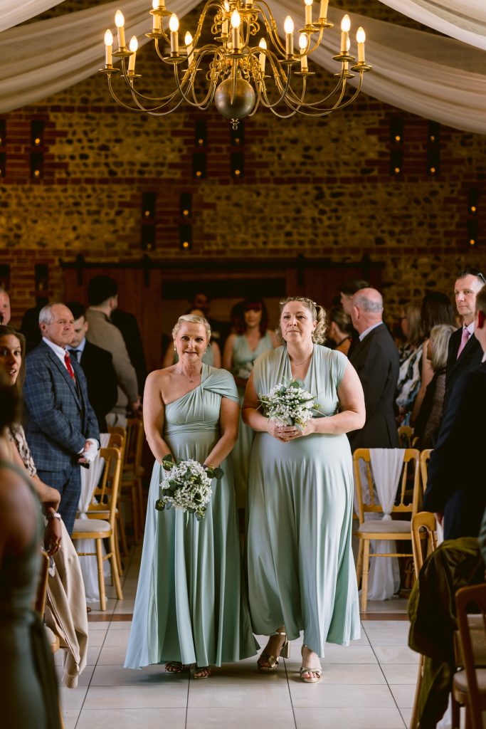 Two bridesmaids in light green dresses walk down the aisle holding bouquets, with guests seated on either side in a beautifully decorated indoor venue—perfect for capturing special moments in Upwaltham Barns Wedding Photography.