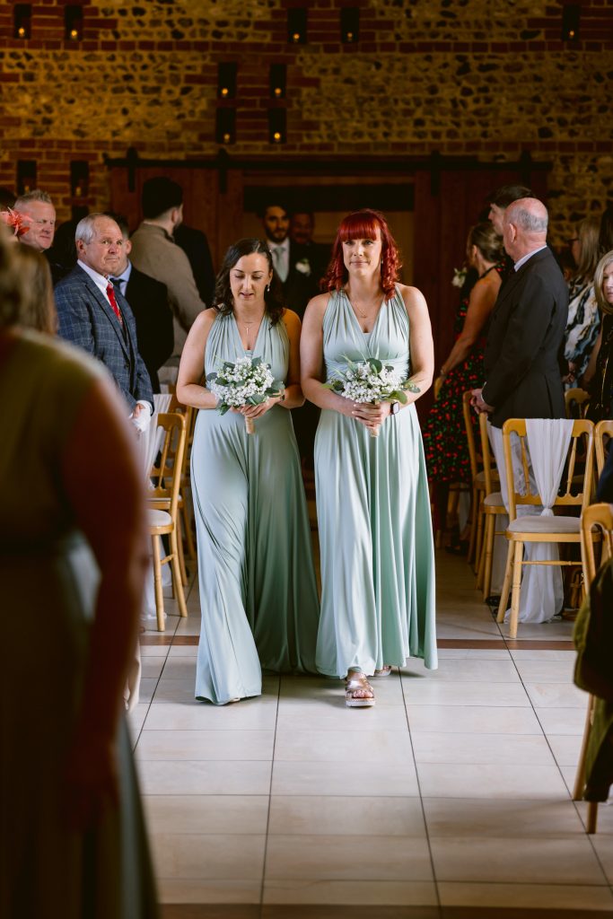 Two bridesmaids in light blue dresses walk down the aisle holding bouquets, with seated guests on either side in a warmly lit venue—capturing a beautiful moment of Upwaltham Barns Wedding Photography.
