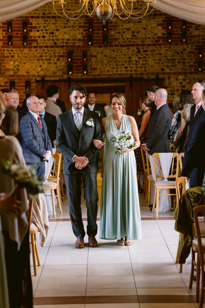 A man in a suit and a woman in a light blue dress walk down the aisle together at an Upwaltham Barns wedding, with guests seated on either side, capturing a perfect moment for wedding photography.