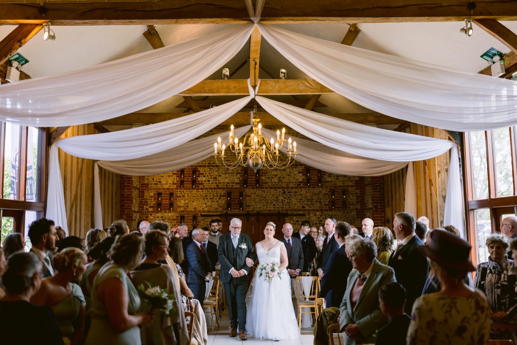A bride in a white dress walks down the aisle with two people at her side in a beautifully decorated indoor venue, as guests look on—capturing the essence of Upwaltham Barns wedding photography.