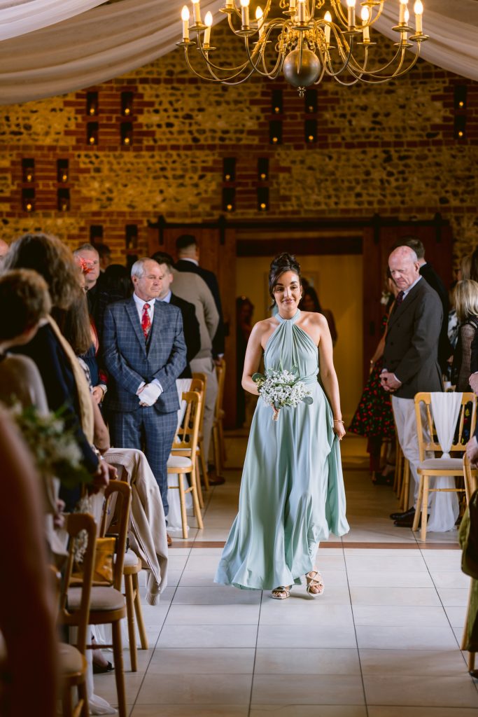 A bridesmaid in a light blue dress walks down the aisle holding a bouquet, with seated guests watching in a warmly lit indoor venue—perfect for timeless Upwaltham Barns Wedding Photography.