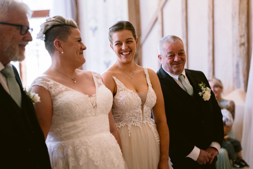 Two brides in white dresses stand side by side, smiling, with two men in suits beside them during an Upwaltham Barns Wedding Photography session indoors.