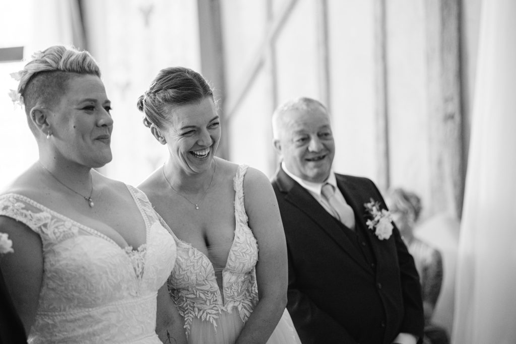 Two women in wedding dresses and a man in a suit stand together indoors, all smiling and looking towards something out of frame—a joyful moment captured through Upwaltham Barns Wedding Photography.