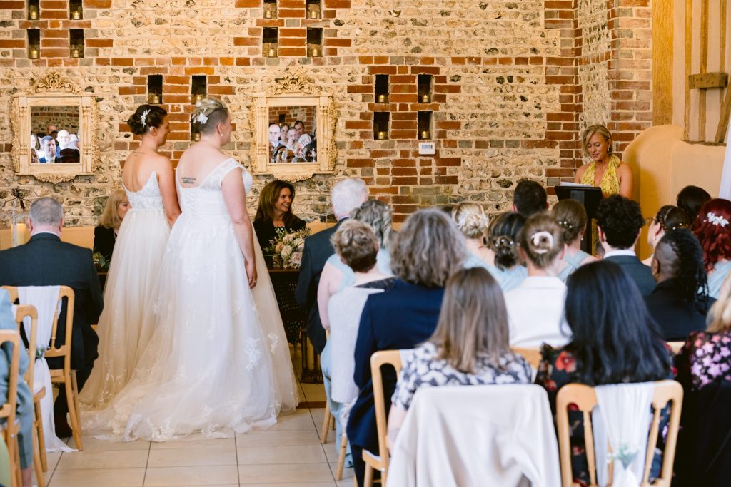Two brides in white dresses stand at the altar facing an officiant, whilst guests watch the ceremony in a brick-walled venue—capturing the romance of Upwaltham Barns wedding photography.