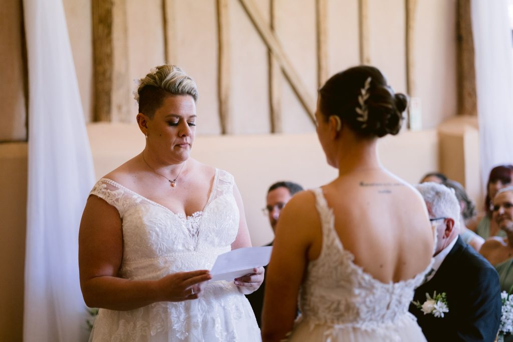 Two brides in white dresses stand facing each other during their Upwaltham Barns Wedding Photography session; one is reading from a piece of paper whilst guests watch in the background.