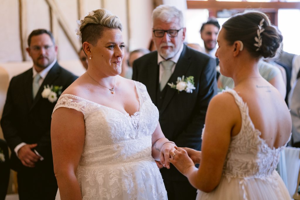 Two women in white wedding dresses hold hands during a wedding ceremony at Upwaltham Barns, surrounded by guests in formal attire—a moment beautifully captured in Upwaltham Barns wedding photography.