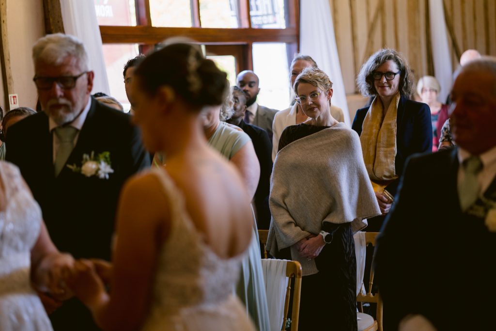 Guests are seated and standing at a wedding ceremony, some watching attentively as the couple hold hands near the front—a beautiful moment captured by Upwaltham Barns Wedding Photography.