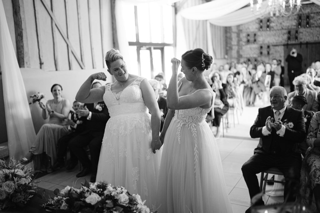 Two brides in wedding dresses stand at the altar, smiling and flexing their arms, as guests watch and clap in a beautifully decorated venue—perfectly capturing the joy of Upwaltham Barns Wedding Photography.