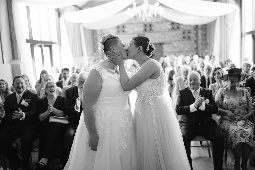 Two brides in wedding dresses kiss at the altar in front of a cheering audience during a wedding ceremony in a beautifully decorated hall, captured in stunning Upwaltham Barns Wedding Photography.