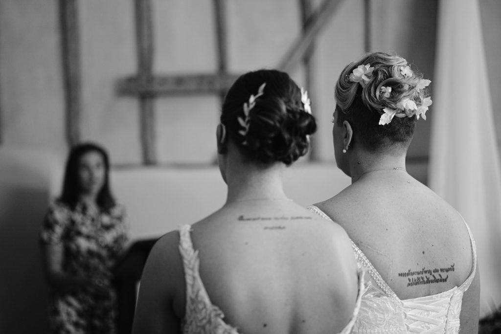 Two women with floral hairpieces and back tattoos, dressed in wedding attire, stand side by side facing an officiant indoors—captured beautifully in this Upwaltham Barns Wedding Photography moment.