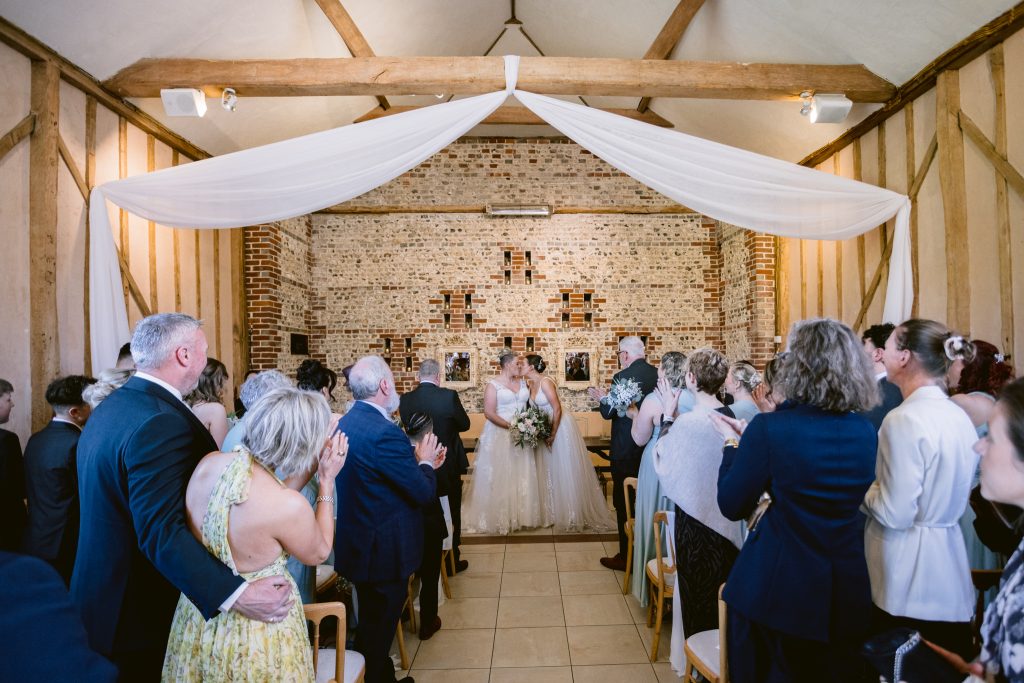 A newlywed couple stands at the front of a rustic indoor venue while guests in formal attire stand and applaud—capturing the magic of the moment with Upwaltham Barns Wedding Photography.