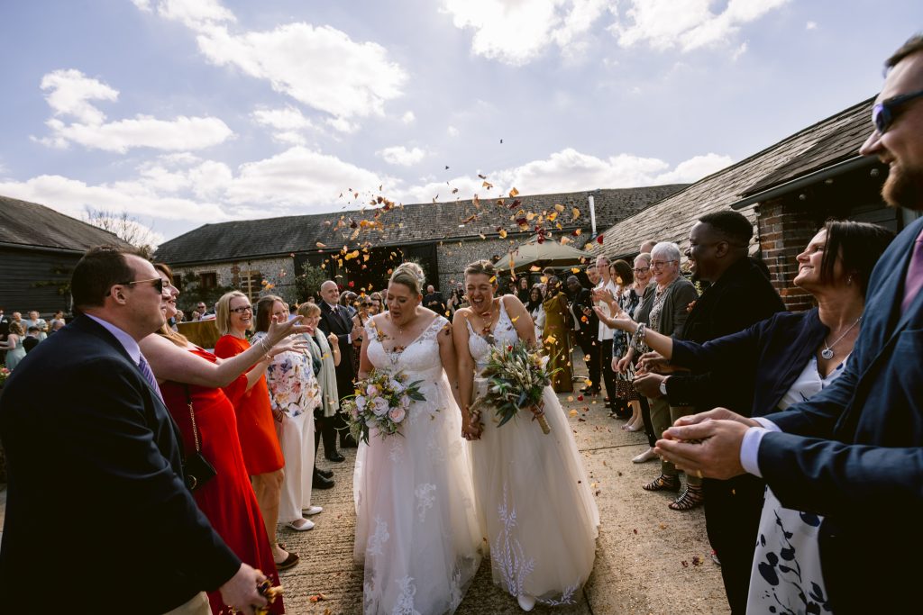 Two brides in wedding dresses walk down an outdoor aisle at Upwaltham Barns, holding hands and bouquets, as guests throw confetti and celebrate under a partly cloudy sky.