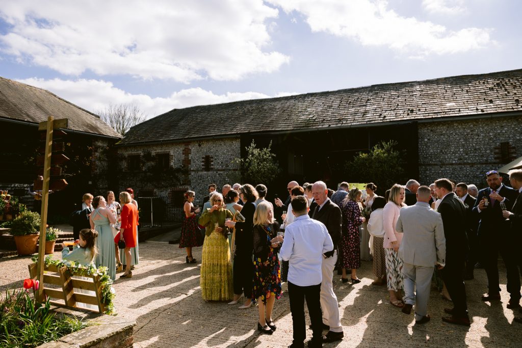A group of people dressed in formal attire gather outdoors in a sunlit courtyard with rustic buildings in the background, capturing the charm of Upwaltham Barns wedding photography.
