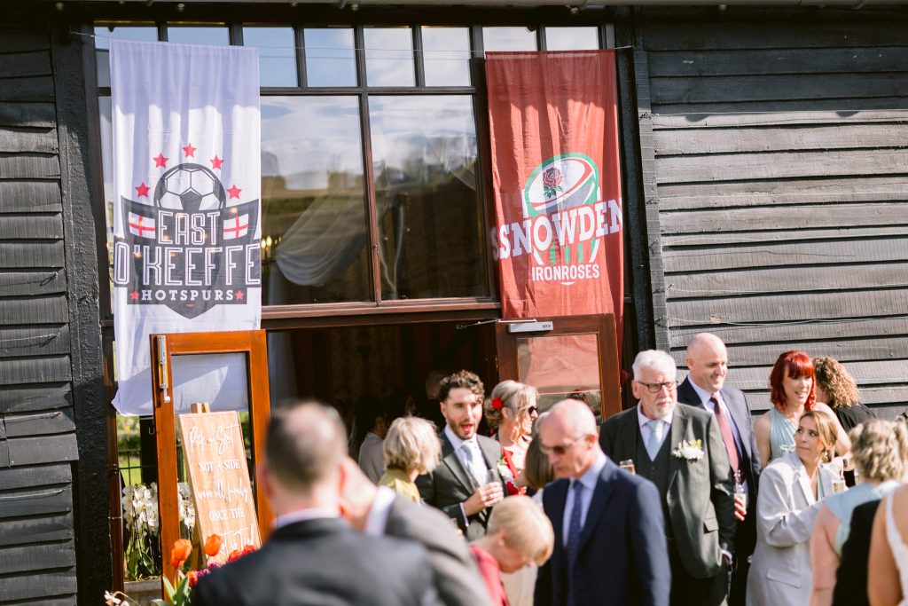 A group of people in formal attire gather outside a black wooden building with two sports banners above the entrance, capturing a moment reminiscent of Upwaltham Barns Wedding Photography.
