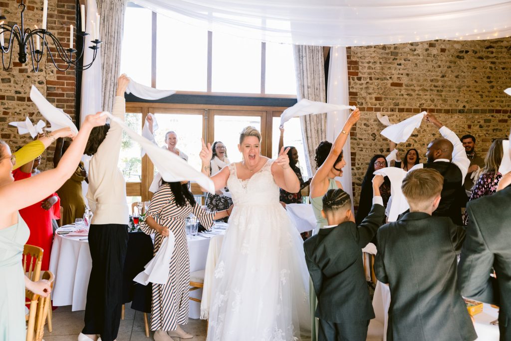 A bride in a white dress stands smiling among guests waving serviettes in the air at a wedding reception, beautifully captured at a bright, rustic venue—perfect for Upwaltham Barns Wedding Photography.