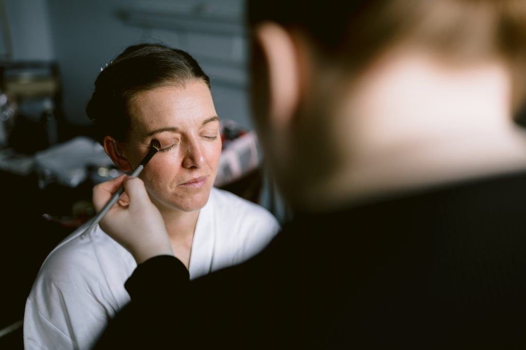A person with closed eyes is having eyeshadow applied by another using a make-up brush, capturing a candid moment perfect for Upwaltham Barns Wedding Photography.