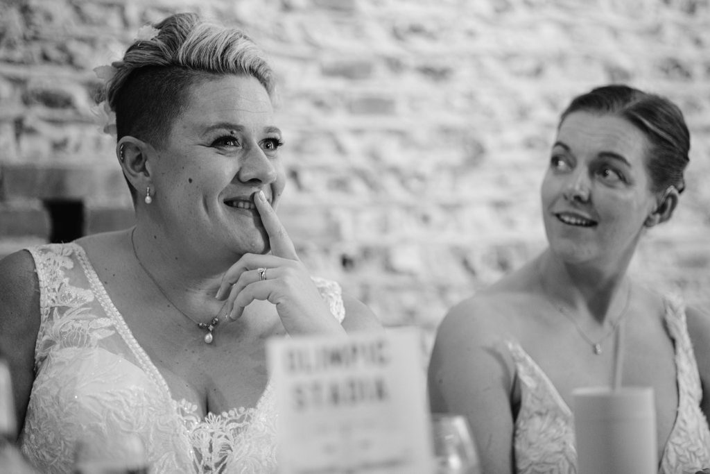 Two women in white dresses sit together at a table, one smiling with her finger to her lips whilst the other looks at her—a charming moment captured by Upwaltham Barns Wedding Photography, with a sign and glass visible in the foreground.