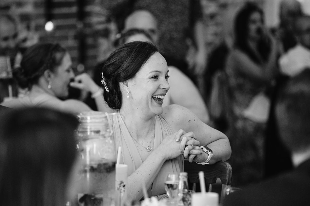 A woman in formal attire sits at a table, smiling and laughing, surrounded by others at what appears to be a social gathering—capturing the joyful moments typical of Upwaltham Barns Wedding Photography.
