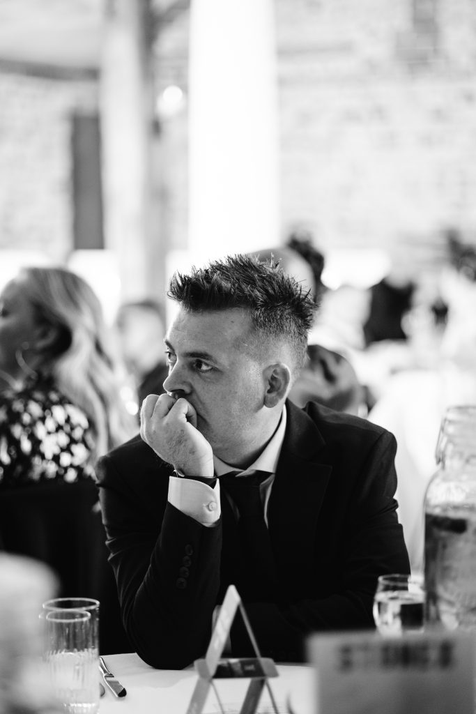 A man in a suit sits at a table with his hand on his chin, looking thoughtful during a formal event—perfectly capturing the candid elegance often seen in Upwaltham Barns Wedding Photography. Other guests are seated in the background.