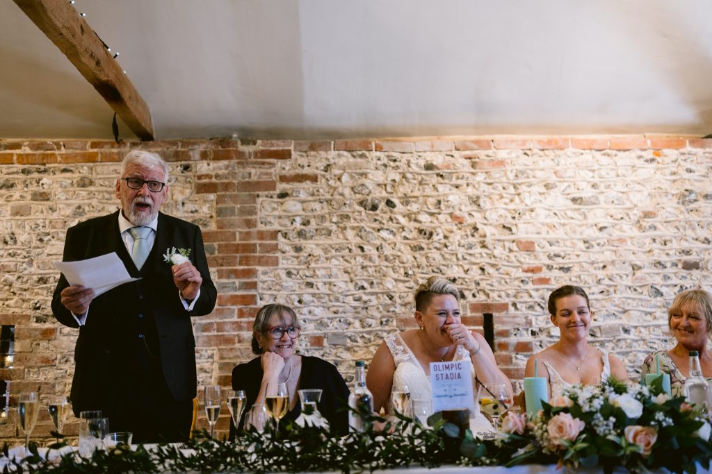 An older man in a suit stands and speaks holding a paper, while four women in formal attire sit at a decorated table, smiling and listening—capturing the warmth of this moment with Upwaltham Barns Wedding Photography.