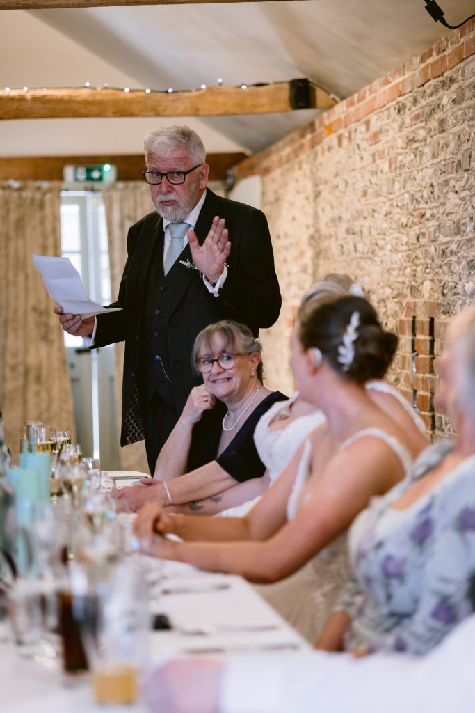 An older man in a suit is standing and giving a speech to seated guests at a long table during a formal event, beautifully captured with Upwaltham Barns Wedding Photography in a charming rustic venue.