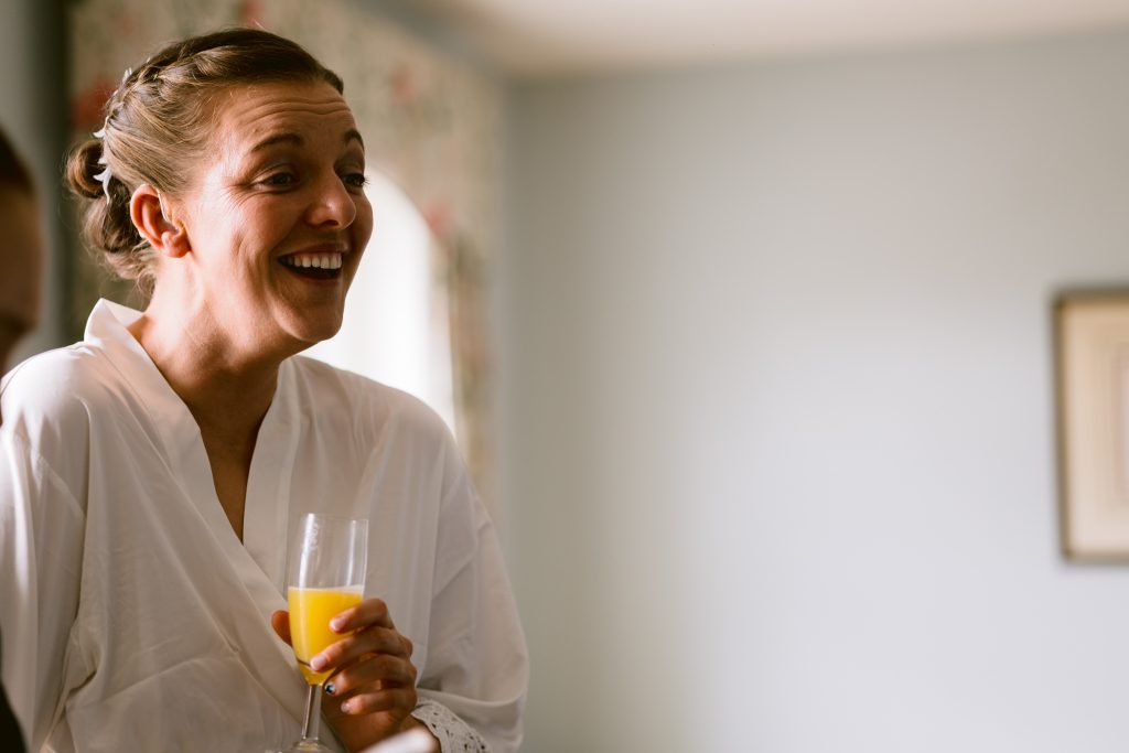 A woman in a white dressing gown smiles whilst holding a glass of orange juice in a softly lit room, capturing the calm anticipation often seen in Upwaltham Barns Wedding Photography.