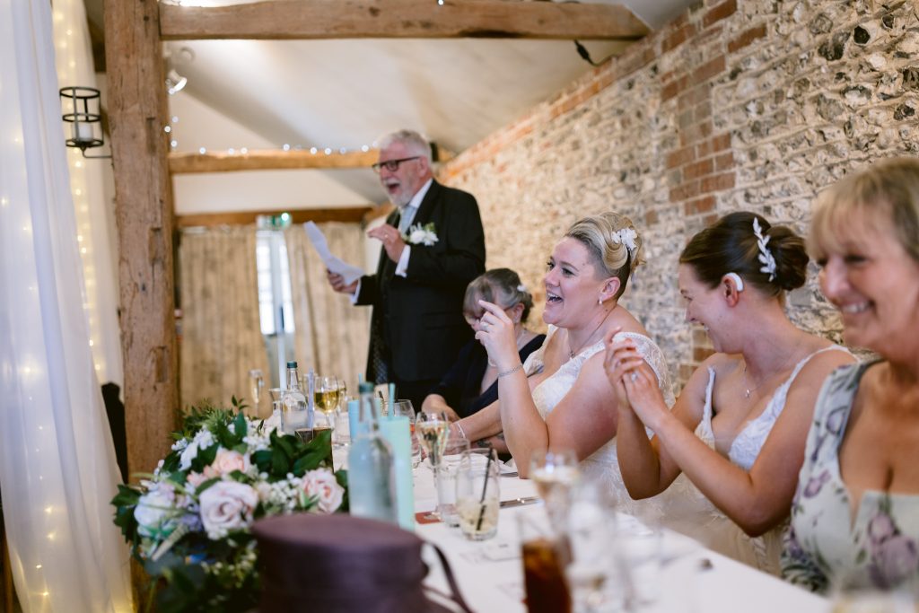 A man gives a speech at a wedding reception table as guests, including the bride, sit and laugh. The rustic brick walls and wooden beams capture the charm of Upwaltham Barns wedding photography.