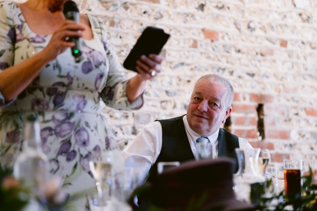 A woman holding a microphone and reading from a device stands next to a seated man in a waistcoat and tie, who looks up at her, against a brick wall background—capturing a candid moment of Upwaltham Barns Wedding Photography.