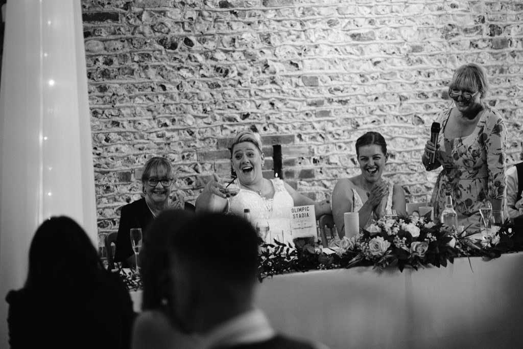 Four women sit at a decorated table, one holding a microphone and another raising a glass, smiling and interacting at what appears to be an Upwaltham Barns Wedding Photography celebration.