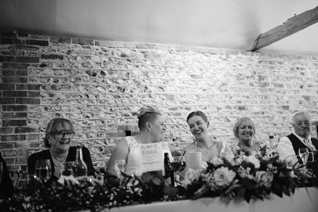 A group of people sit at a decorated table, smiling and talking, in front of a brick wall at what appears to be a wedding reception—capturing joyful moments perfect for Upwaltham Barns Wedding Photography.