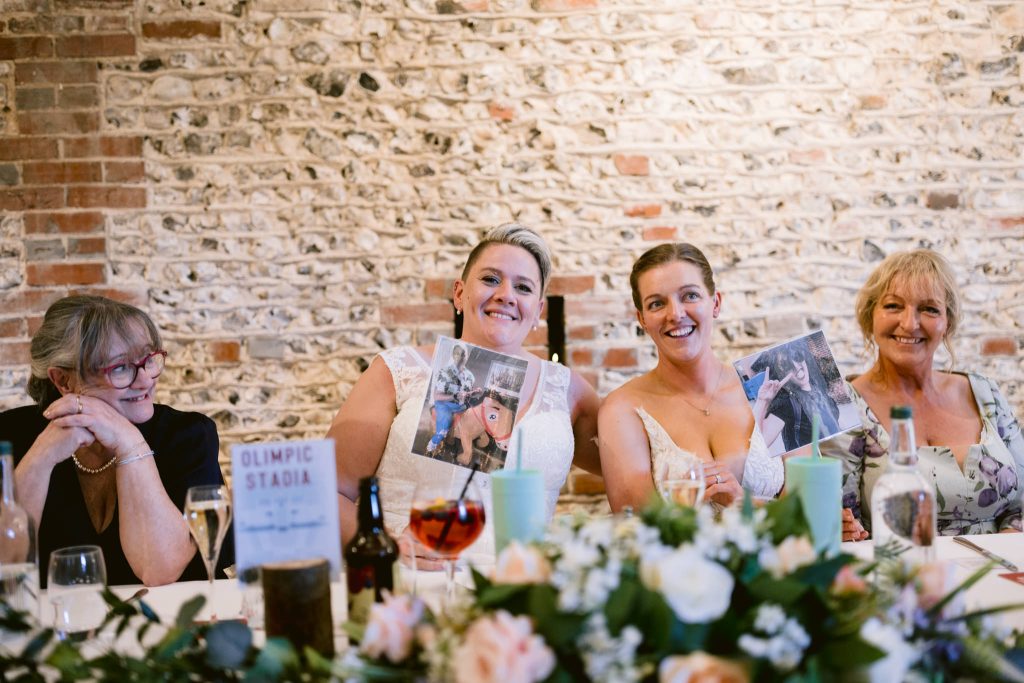 Four women sit at a decorated table, two smiling and holding printed photos. A sign reading "OLIMPIC STADIA" sits on the table, with a brick wall behind them—capturing the joy of an Upwaltham Barns Wedding Photography moment.