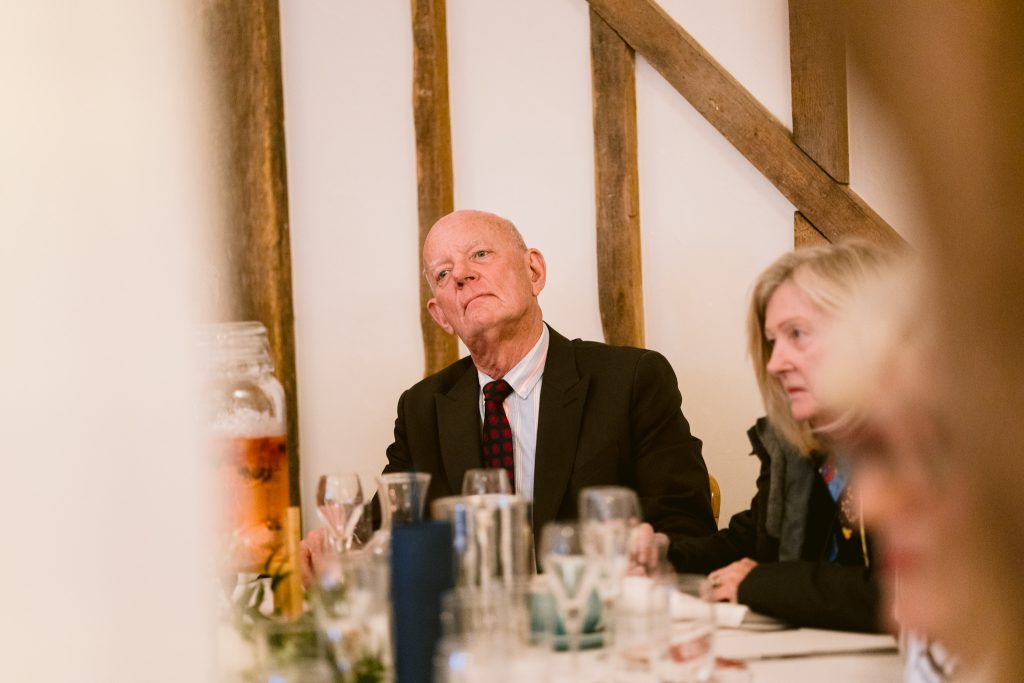 An older man in a suit and a woman sit at a table with empty glasses and tableware, in a room with wooden beams—capturing the warmth of an Upwaltham Barns Wedding Photography moment.