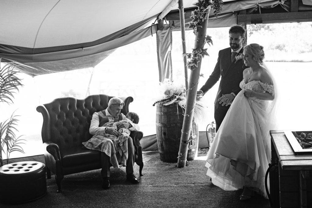 An elderly person sits on a settee holding a baby while a bride and groom stand nearby under a marquee, all dressed formally.