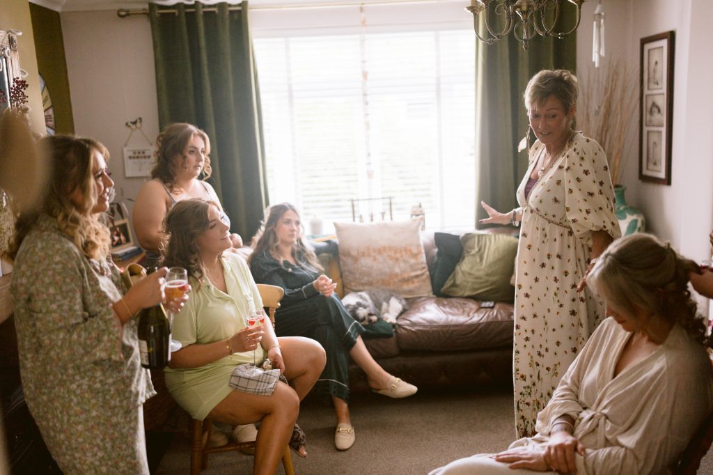 Six women gather in a cosy living room, some seated and some standing, talking and holding drinks near a window with green curtains.