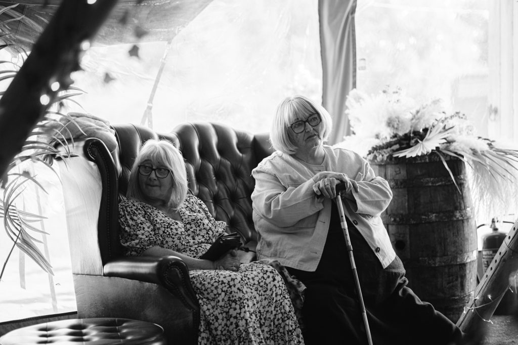 Two older women with glasses sit indoors; one holds a mobile on a sofa, the other sits beside her with a walking stick. Dried flowers and a wooden barrel are in the background.